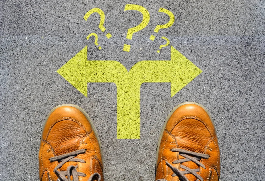 アスファルトに描かれた左右の分岐矢印とはてなマークの前に立つ、茶色の靴を履いた人の足元