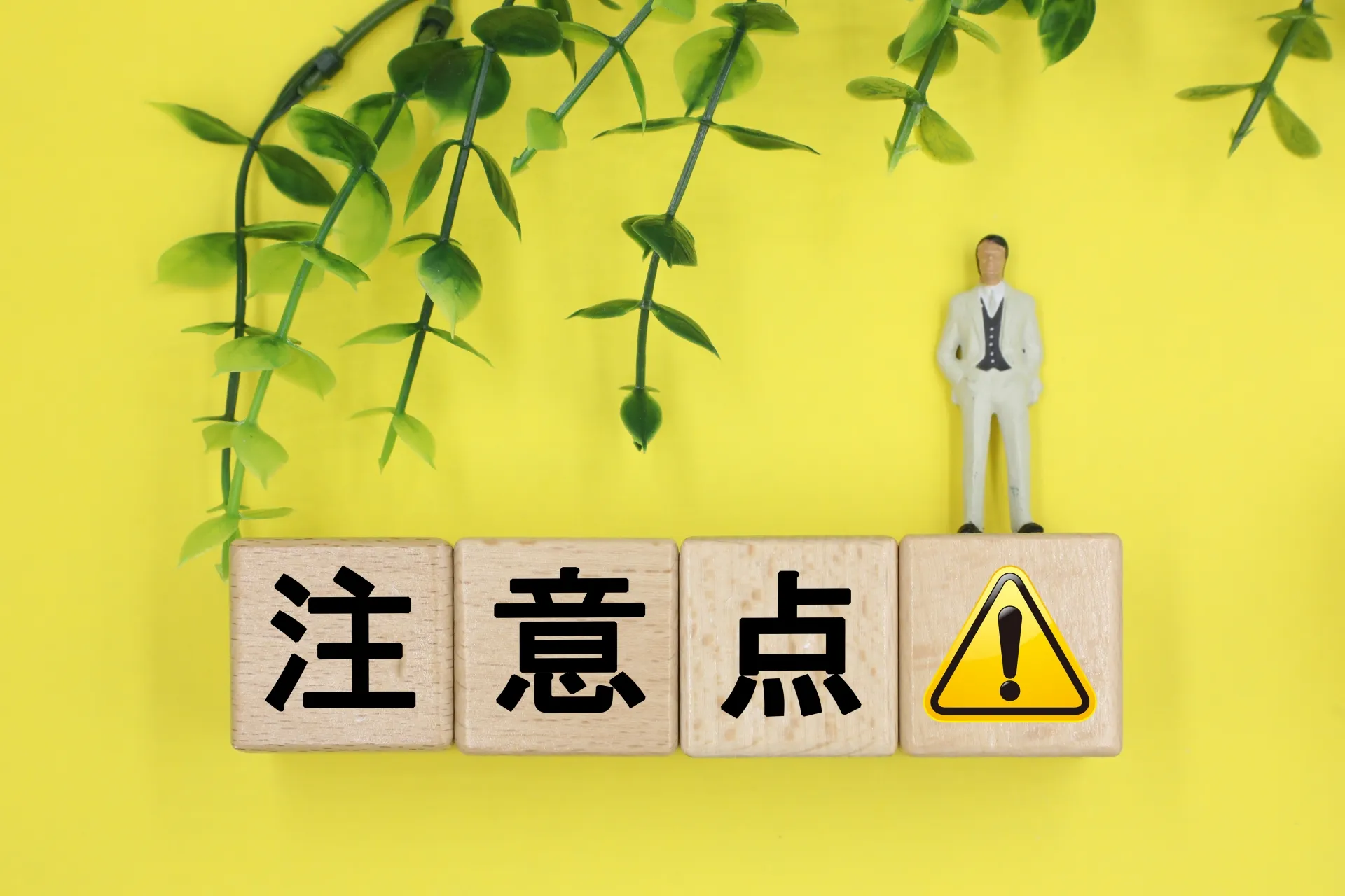 黄色の背景に「注意点」と書かれた木のブロックと警告マークの上に立つ人物模型と植物の装飾