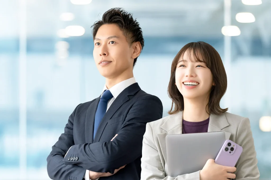 オフィスを背景に、笑顔で前を見据えるスーツ姿の男性とノートパソコンを持つ女性のビジネスパーソン