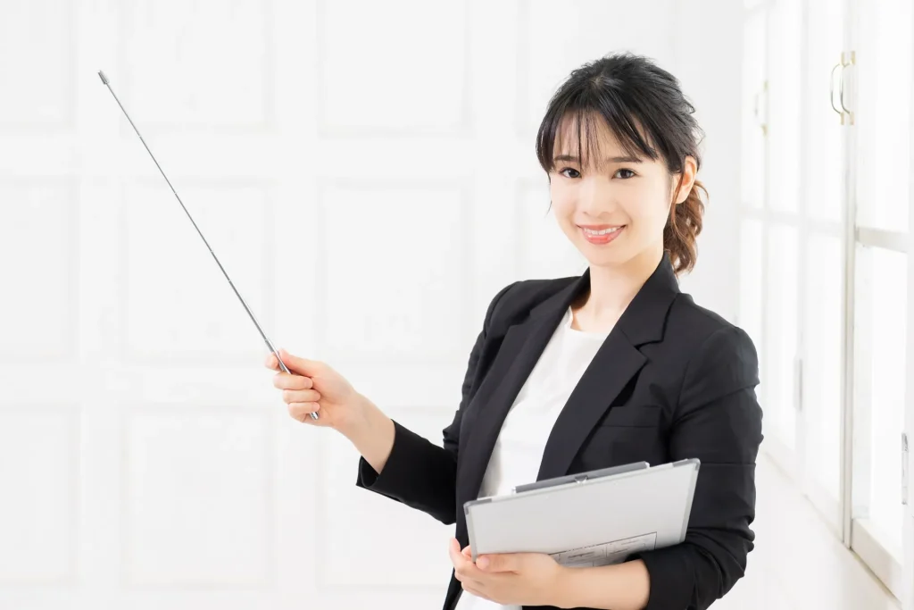 指示棒とバインダーを持ち、笑顔でプレゼンテーションや案内をするスーツ姿の女性