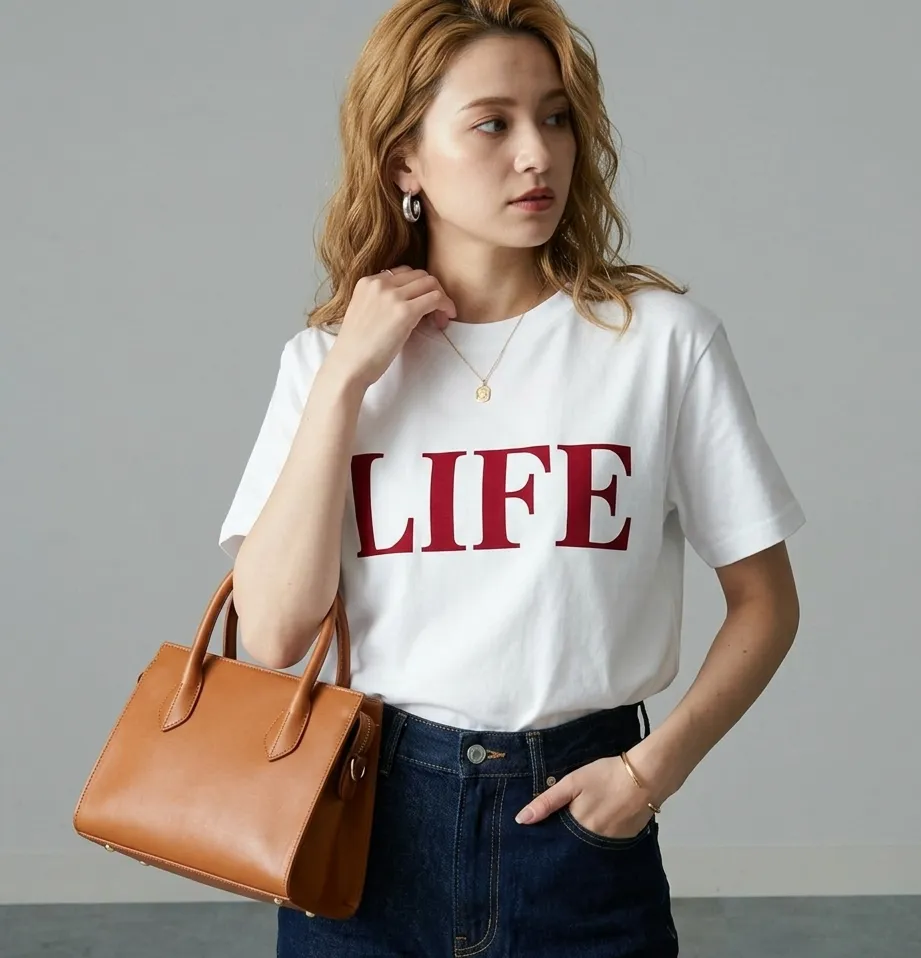 胸元に赤字でLIFEと書かれた白Tシャツに、濃紺のデニムと茶色のレザートートバッグを合わせた女性のコーディネート