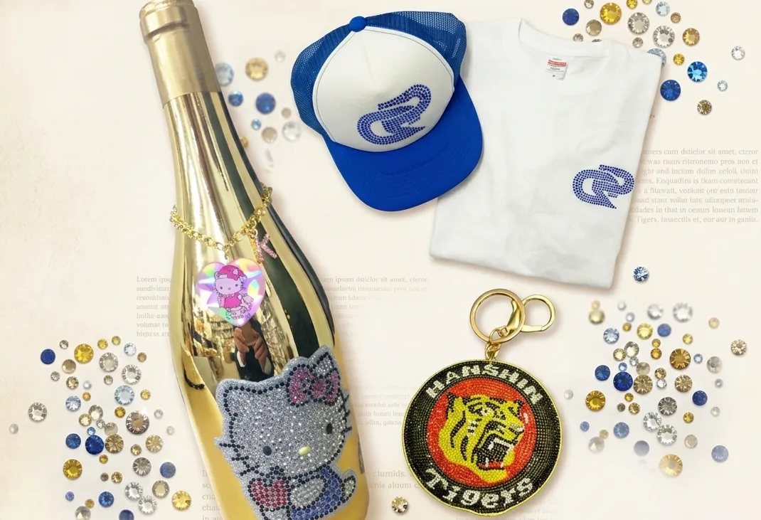 ラインストーンでデコレーションされたTシャツ、キャップ、キーホルダー、ゴールドのボトルが、散りばめられたラインストーンと共に並べられた横長のデザイン画像