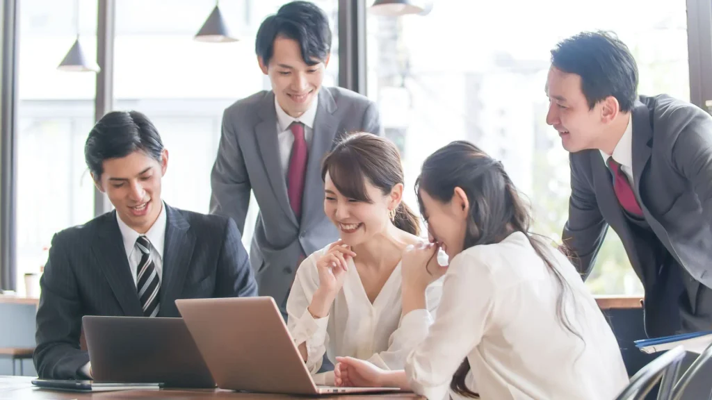 オフィスでノートパソコンを囲みながら笑顔で話し合う5人のビジネスチーム