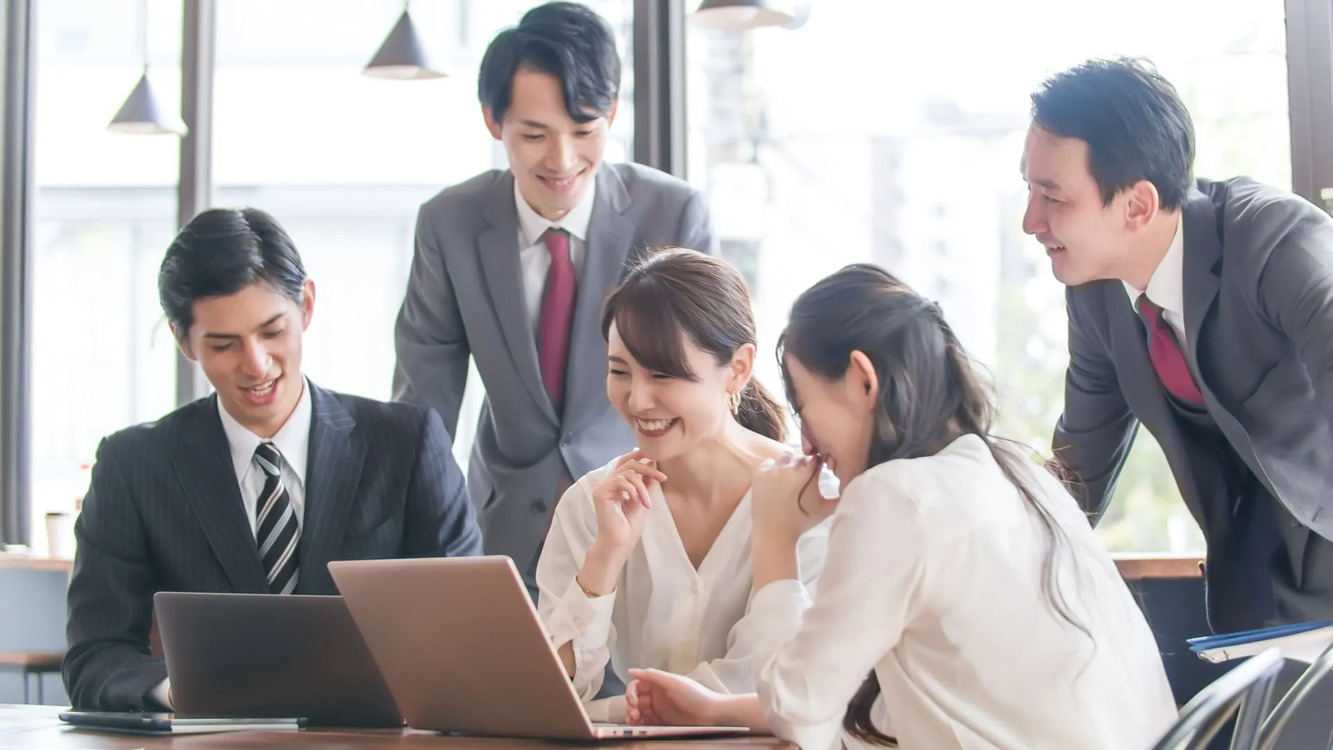 オフィスでノートパソコンを囲みながら笑顔で話し合う5人のビジネスチーム